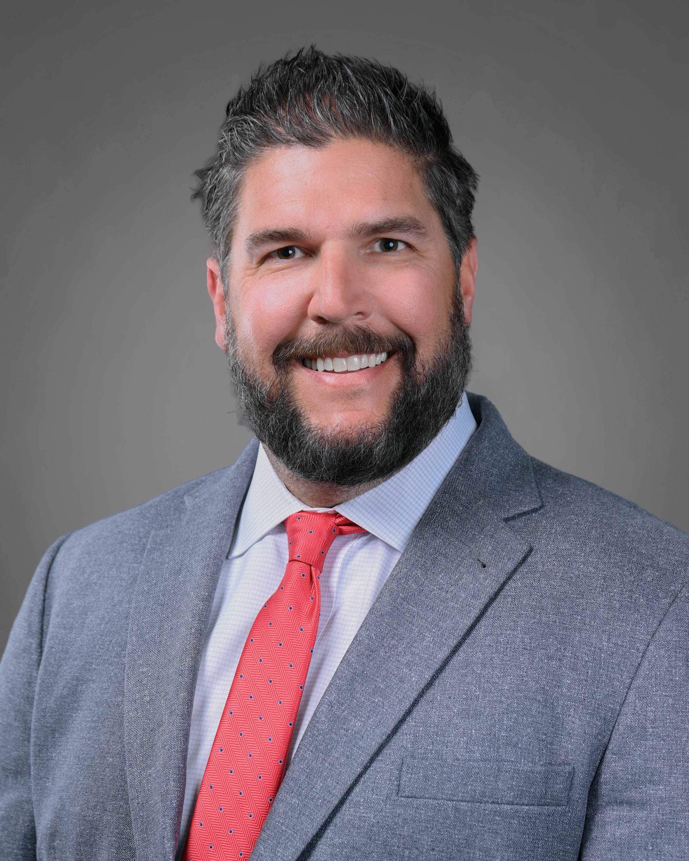 Professional headshot of a smiling man with a beard, wearing a light gray suit, white dress shirt, and coral-colored tie against a gray studio background.